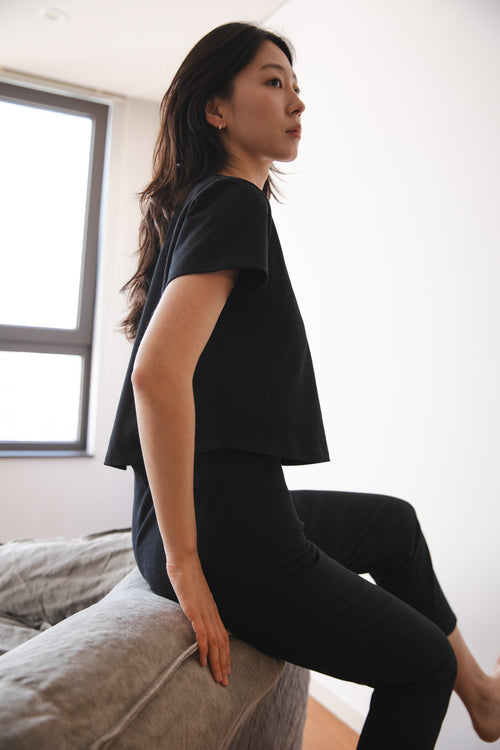 A woman wearing a black short-sleeve crop top and black pants sits on the edge of a gray sofa in a bright room, shown in profile.